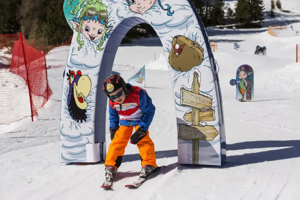 Kids on ski fahren durch einen bunt gestalteten Torbogen im Schnee während eines gratis Skikurs für Kinder, umgeben von fröhlichen Tier- und Fantasiefiguren.