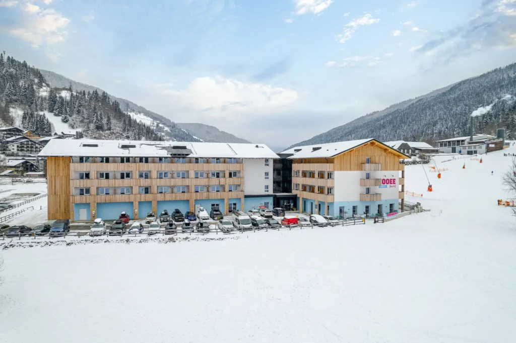 Das COOEE alpin Hotel Bad Kleinkirchheim von außen im Winter. Rechts vom Hotel ist eine Skipiste und hinter dem Hotel sieht man verschneite, mit Wäldern bedeckte Berge.