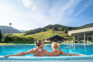 Two people relaxing at the outdoor pool of the thermal spa Römerbad in summer with a view of the cable car Kaiserburgbahn.