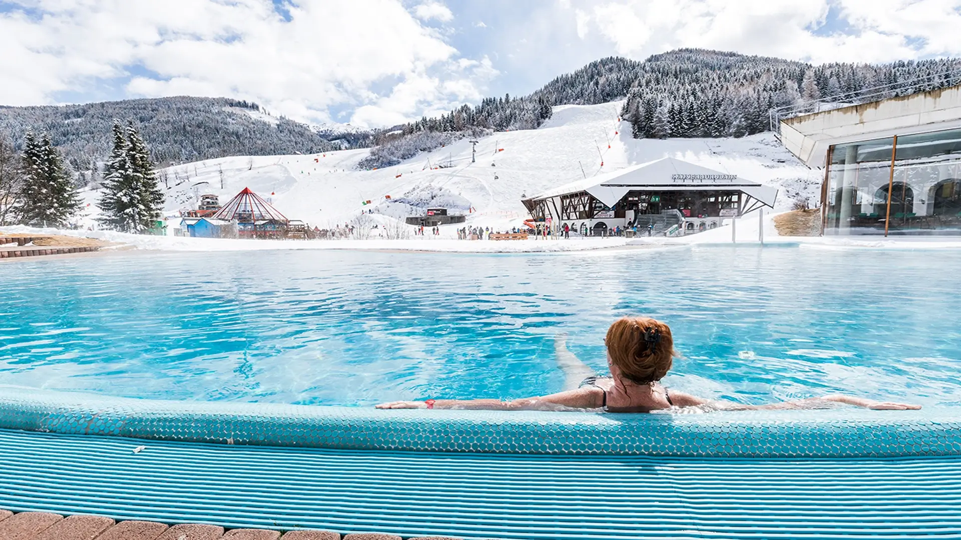 Eine Person mit roten Haaren entspannt sich in einem Infinity-Pool im Thermal Römerbad in Bad Kleinkirchheim und blickt auf ein verschneites Skigebiet mit Bergen im Hintergrund. In der Ferne sind Skifahrer und schneebedeckte immergrüne Bäume zu sehen. Der Himmel ist teilweise bewölkt, was eine ruhige und malerische Winterlandschaft schafft.