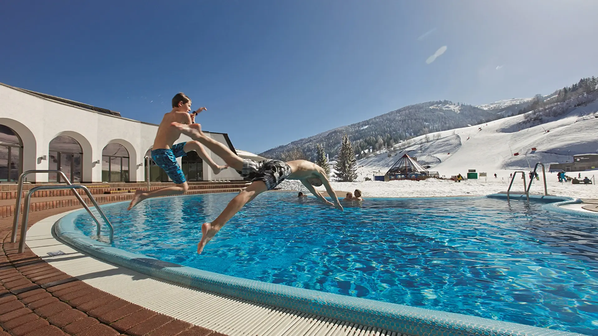 Zwei Jugendliche springen in den Outdoor Pool im Thermal Römerbad im Winter bei Sonnenschein. Im Hintergrund kann man die Piste erkennen.