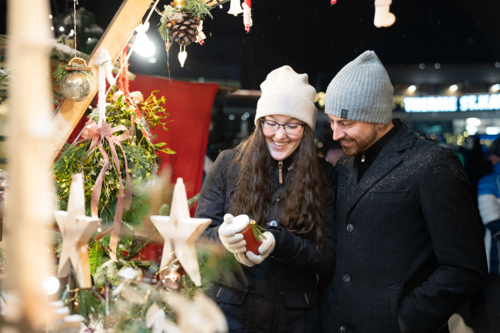 Ein Mann und eine Frau besuchen einen Adventsmarkt und schauen sich bei einem Stand rote, festlich dekorierte Marmelade an, welche die Frau in der Hand hält. Sie tragen dicke Jacken und Hauben.