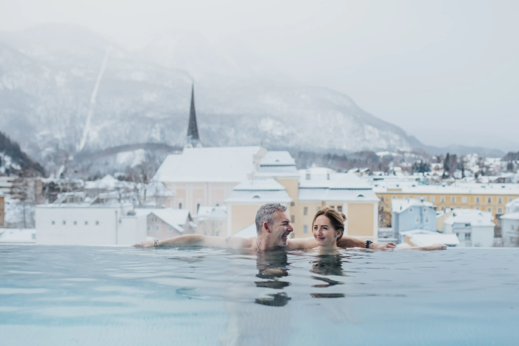 Zwei Personen relaxen in einem Außenpool mit Blick auf das verschneite Bad Ischl mit der Kirche und Bergen im Hintergrund.