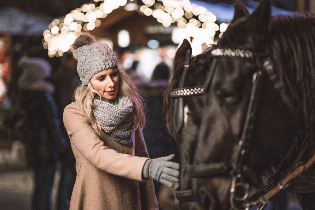 Eine Frau mit einem Mantel, Schal, Haube und Handschuhen streichelt ein schwarzes Pferd, das vor eine Kutsche gespannt ist. Im Hintergrund erkennt man den beleuchteten Stand auf einem Adventmarkt und Leute stehen davor.