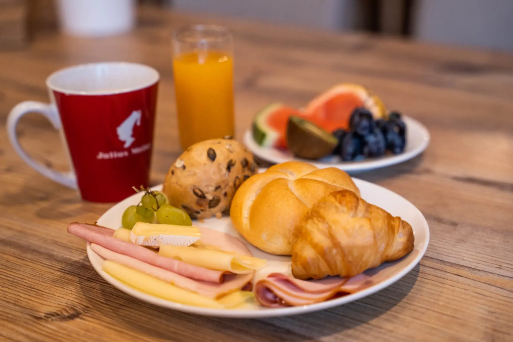 Frühstücksteller mit einem Croissant, einer Semmel, Wurst- und Käseaufschnitt und Obst. Ein Kaffee und ein Orangensaft stehen auch auf dem Tisch.