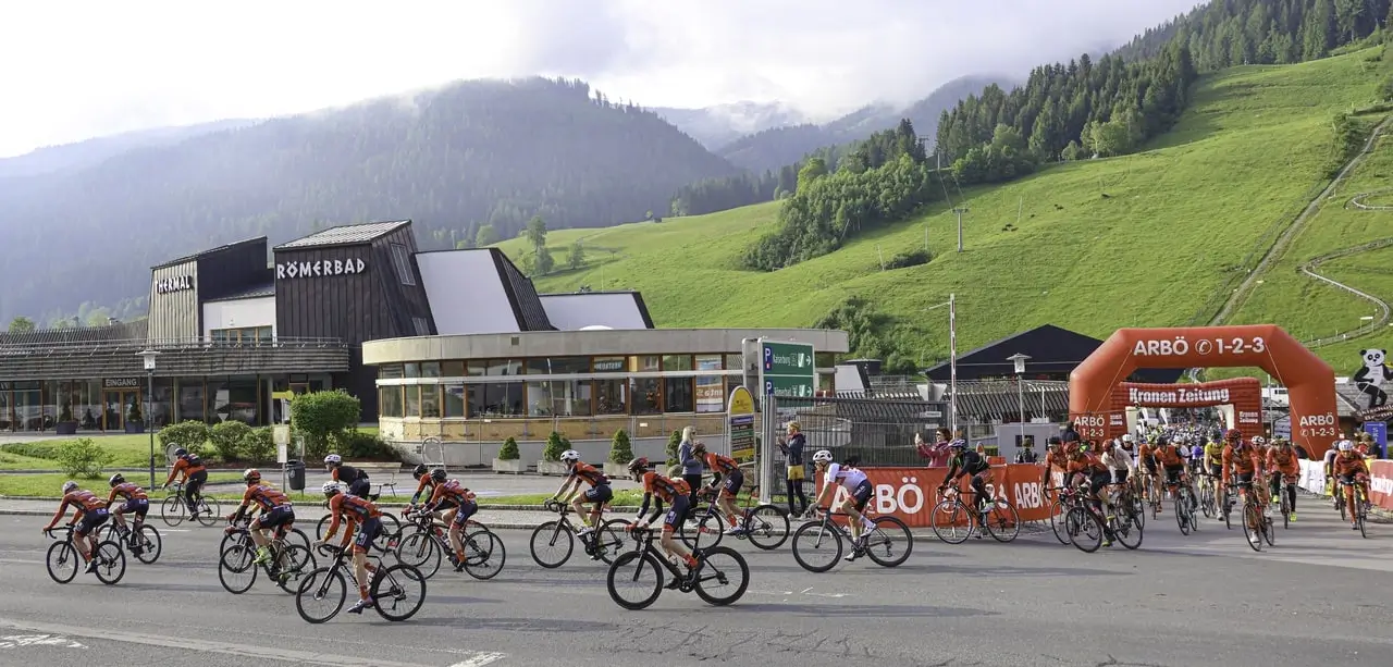 Der Start des ARBÖ Radmarathons in Bad Kleinkirchheim mit vielen teilnehmenden Personen die gerade aus der Start Zone mit dem roten Bogen, der mit Luft aufgeblasen wird, hinaus fahren. Der Startbereich liegt direkt neben dem Thermal Römerbad und der Kaiserburgbahn.