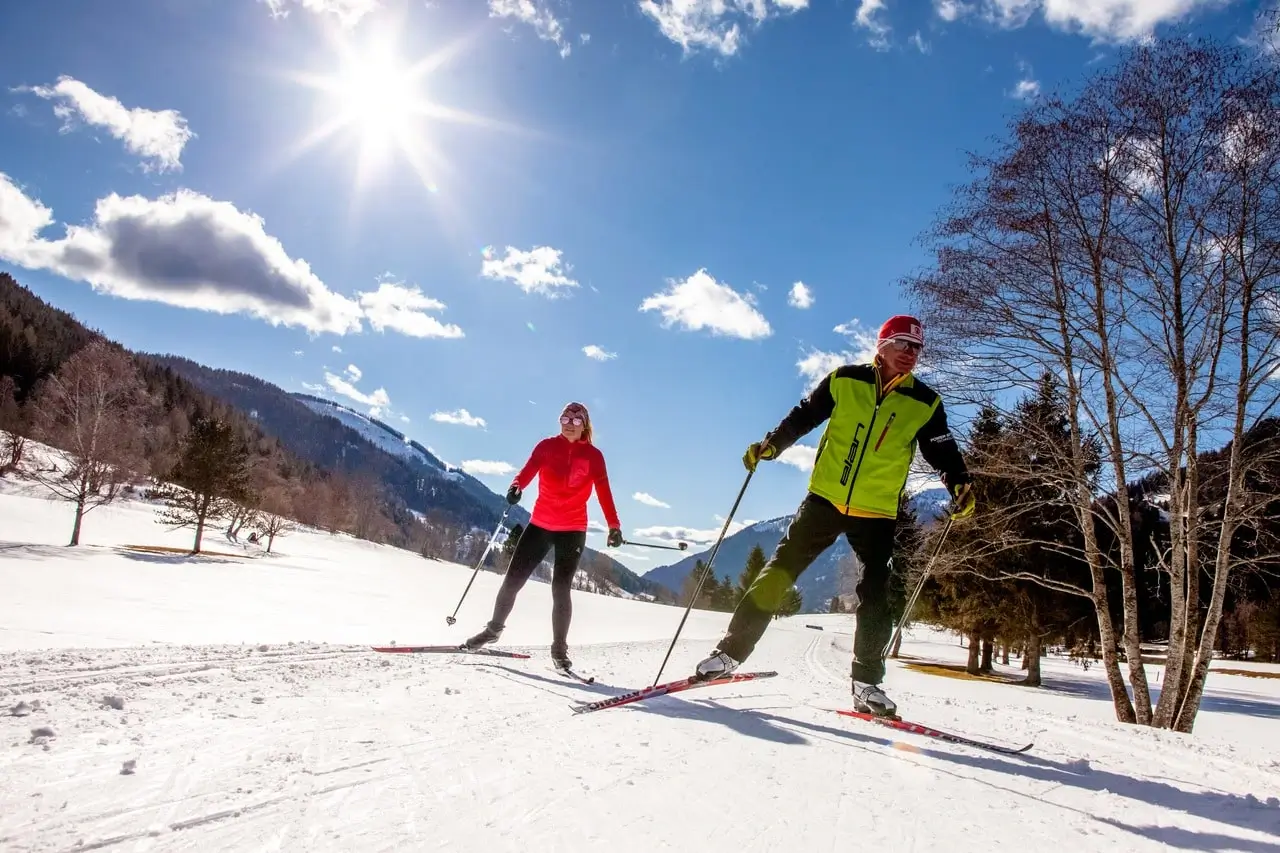 Zwei Personen mit Langlaufskiern skaten auf einer Loipe in einem verschneiten Tal. Die Sonne scheint vom Himmel herab auf die umliegenden Wälder und Berge.