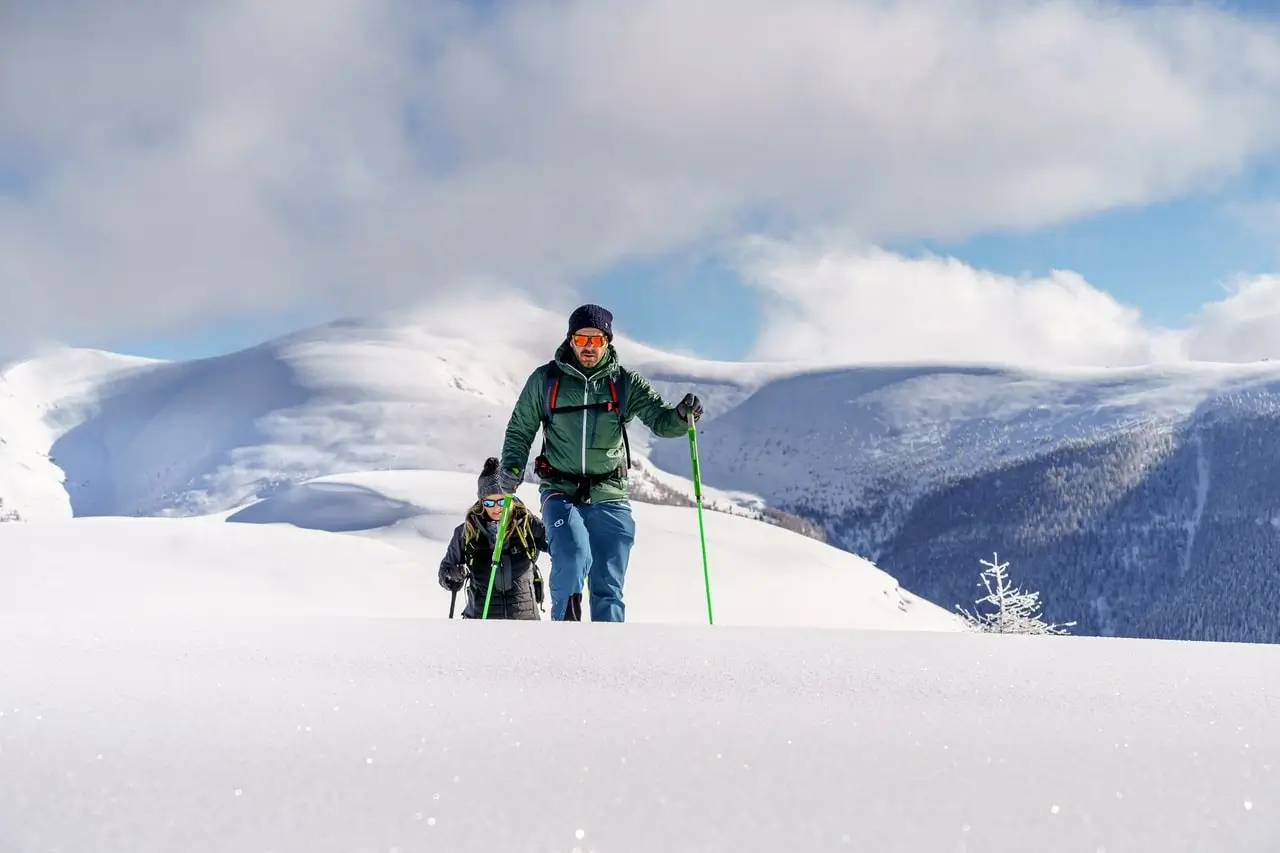 Zwei Personen mit Stöcken gehen durch den Tiefschnee auf einem Bergplateau. Hinter ihnen sieht man auf die schneebedeckten Gipfel und der Schnee glänzt in der Sonne.