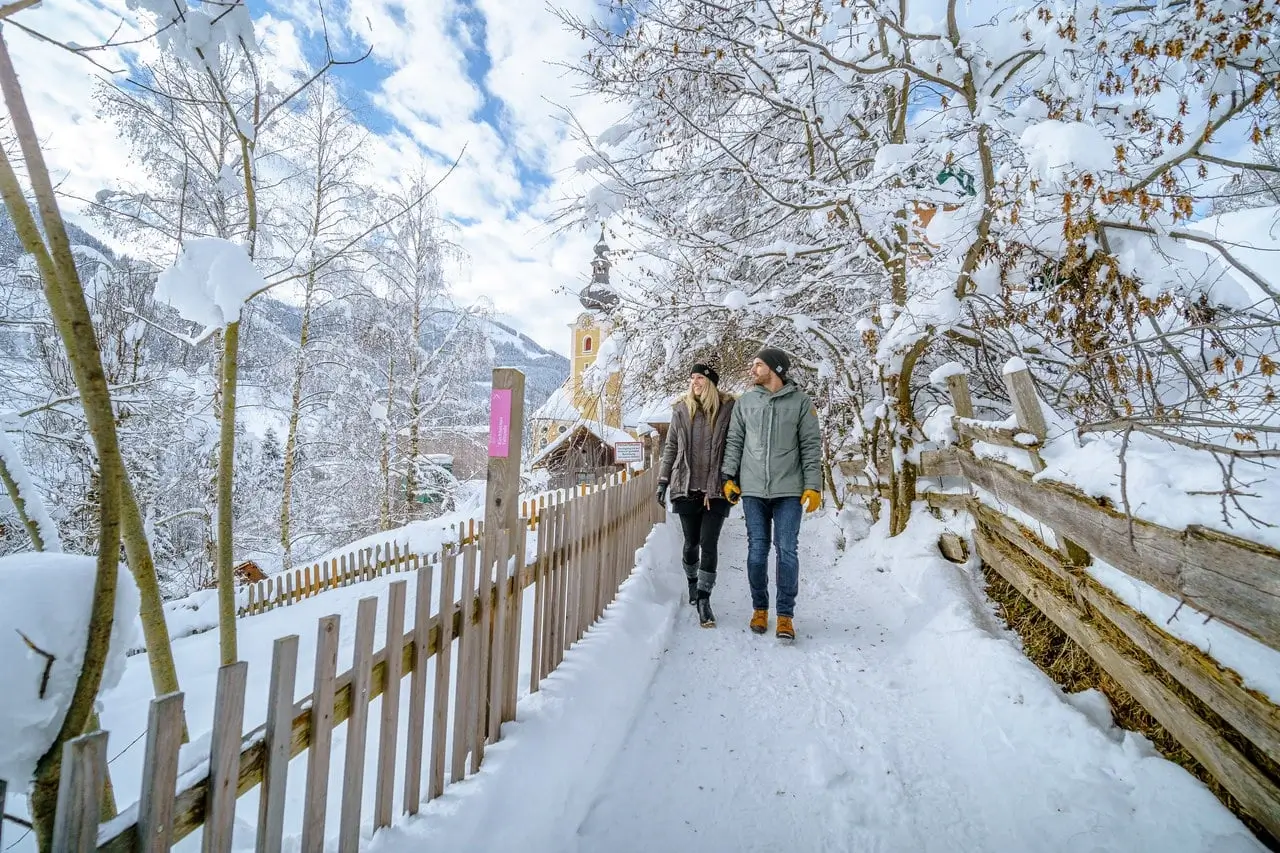 Zwei Personen spazieren einen schneebedeckten, jedoch geräumten Weg entlang. Auf beiden Seiten des Weges stehen Holzzäune und schneebedeckte Bäume. Hinter ihnen ist eine gelbe Kirche und ein kleines Holzhaus zu erkennen.