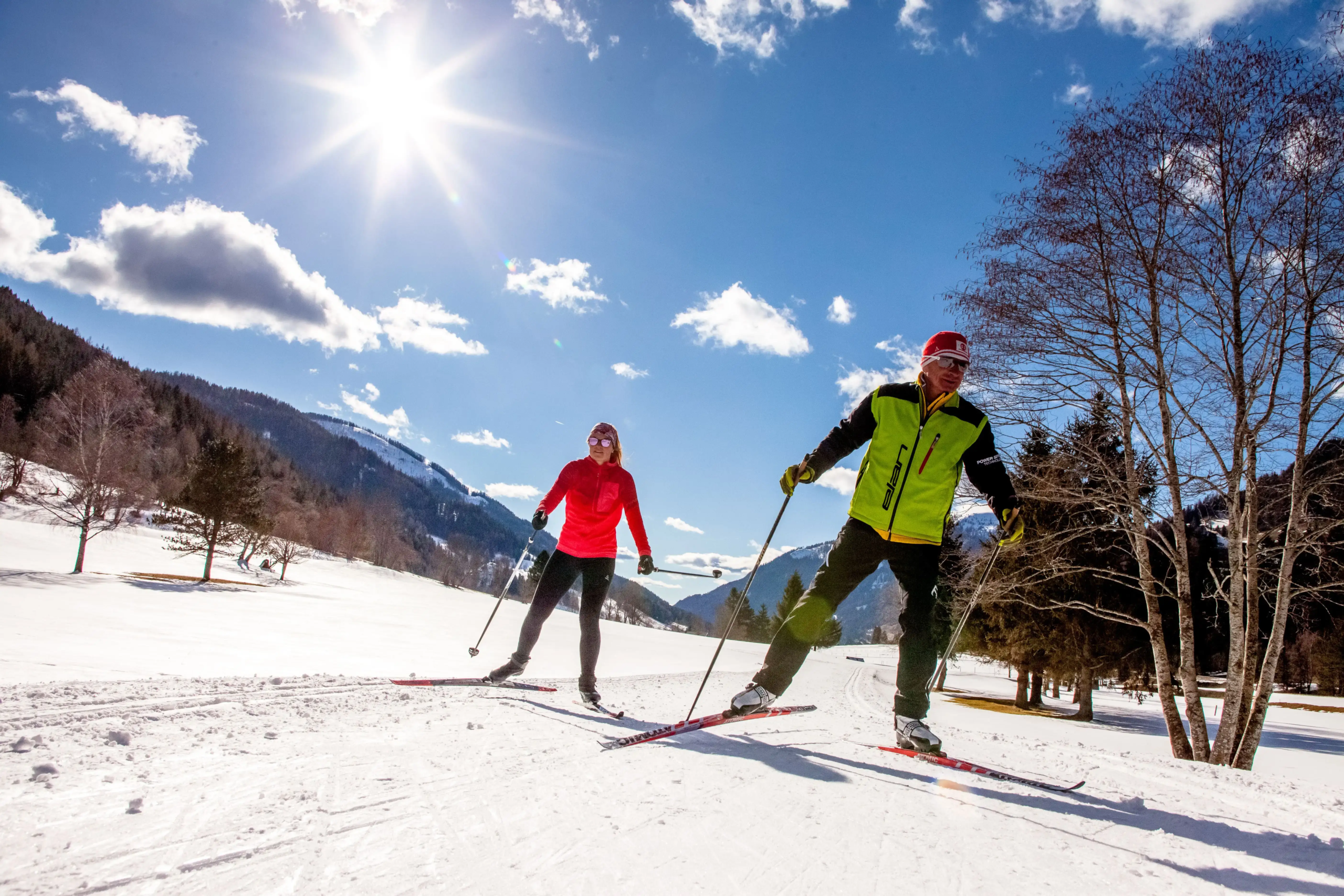 Zwei Personen mit Langlaufskiern skaten auf einer Loipe in einem verschneiten Tal. Die Sonne scheint vom Himmel herab auf die umliegenden Wälder und Berge.