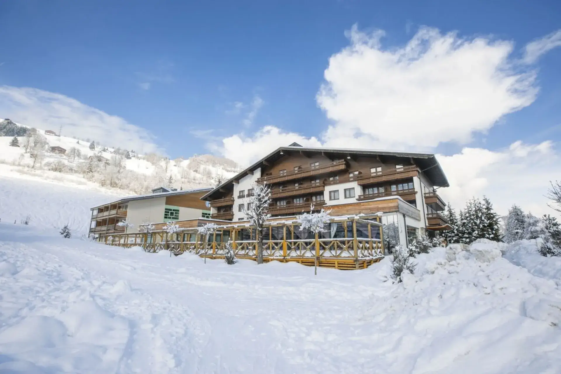 Verschneites COOEE alpin Hotel Kaprun | Zell am See in Kaprun, umgeben von einer Schneelandschaft. Weißes Hotelgebäude mit dunkler Holzverkleidung. Vor der Terrasse eingeschneite Bäume.