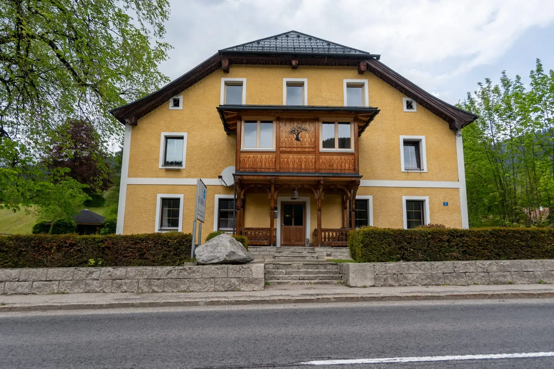 Vista esterna degli Ulmenhof Apartments a Gosau, con la storica facciata gialla, il balcone in legno riccamente decorato e l'ingresso, circondati da alberi rigogliosi.
