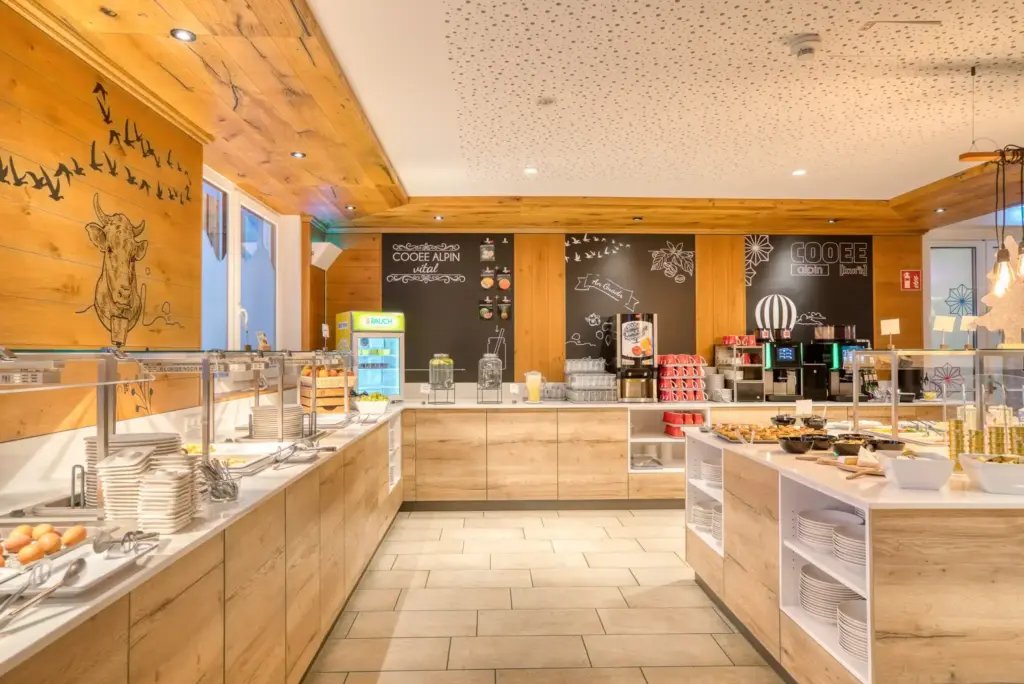 Buffet in einem Hotel. Links sind Eier, ein Obstkorb und Teller. An der Rückwand stehen zwei Kaffeemaschinen, ein Saftspender und zwei Wasserspender. Rechts ist eine Buffetinsel mit Aufstrichen, Wurst- und Käseaufschnitt.