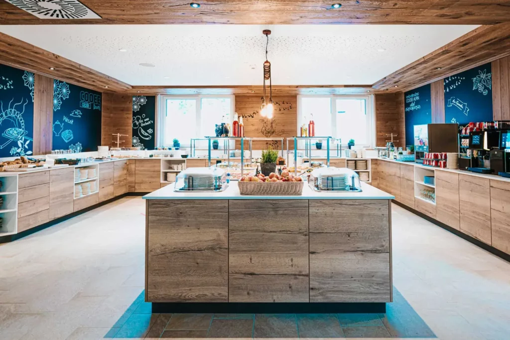 Buffet con rivestimento in legno, dotato di due macchine da caffè, distributori di succhi e un piccolo frigorifero sul lato destro. A sinistra si trovano cestini per il pane, taglieri di legno, sei vasetti di marmellata e quattro distributori di muesli. Al centro della sala si trova un’isola buffet con un cesto di frutta, piatti, affettati e formaggi.