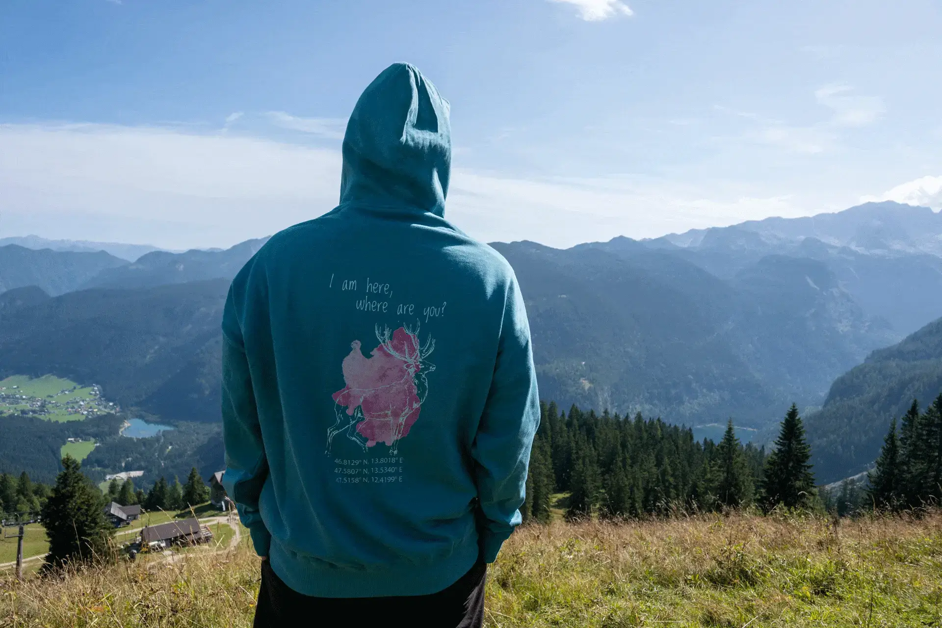 Person mit türkisfarbenem COOEE alpin Hoodie mit Hirsch-Motiv und Aufschrift „I am here, where are you?“ steht auf einer Bergwiese und blickt ins Alpenpanorama – nachhaltige Outdoor-Mode und Merchandise für Urlaub in Österreich.