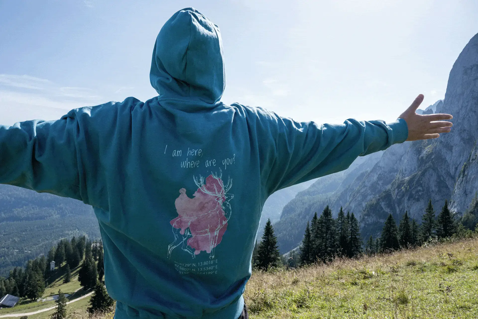 Person mit türkisfarbenem COOEE alpin Hoodie mit Hirsch-Motiv und Aufschrift „I am here, where are you?“ steht mit ausgebreiteten Armen vor der Bergkulisse in den Alpen – nachhaltiger Outdoor-Hoodie für Abenteuer und Naturgenuss in Österreich.