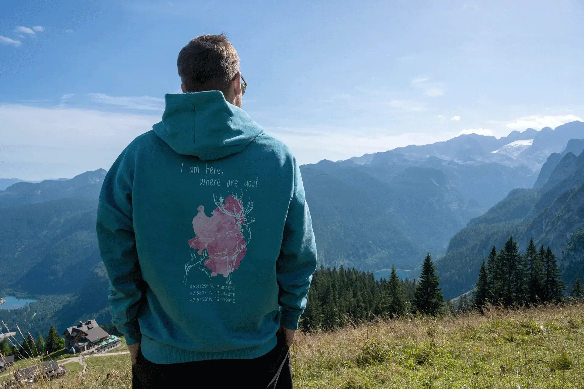 Person mit türkisfarbenem COOEE alpin Hoodie mit Hirsch-Motiv und Aufschrift „I am here, where are you?“ steht vor der beeindruckenden Bergkulisse der Alpen – nachhaltiger Hoodie für Outdoor-Abenteuer, Wandern und Naturgenuss in Österreich.