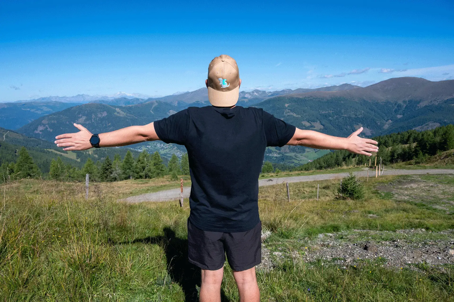 Wanderer mit beige-brauner COOEE alpin Cap „Alinga“ auf einem Berggipfel in Österreich, breitet die Arme aus und genießt die Aussicht auf die Alpen – stylische Outdoor-Kappe für Abenteuer, Wandern und Natur.