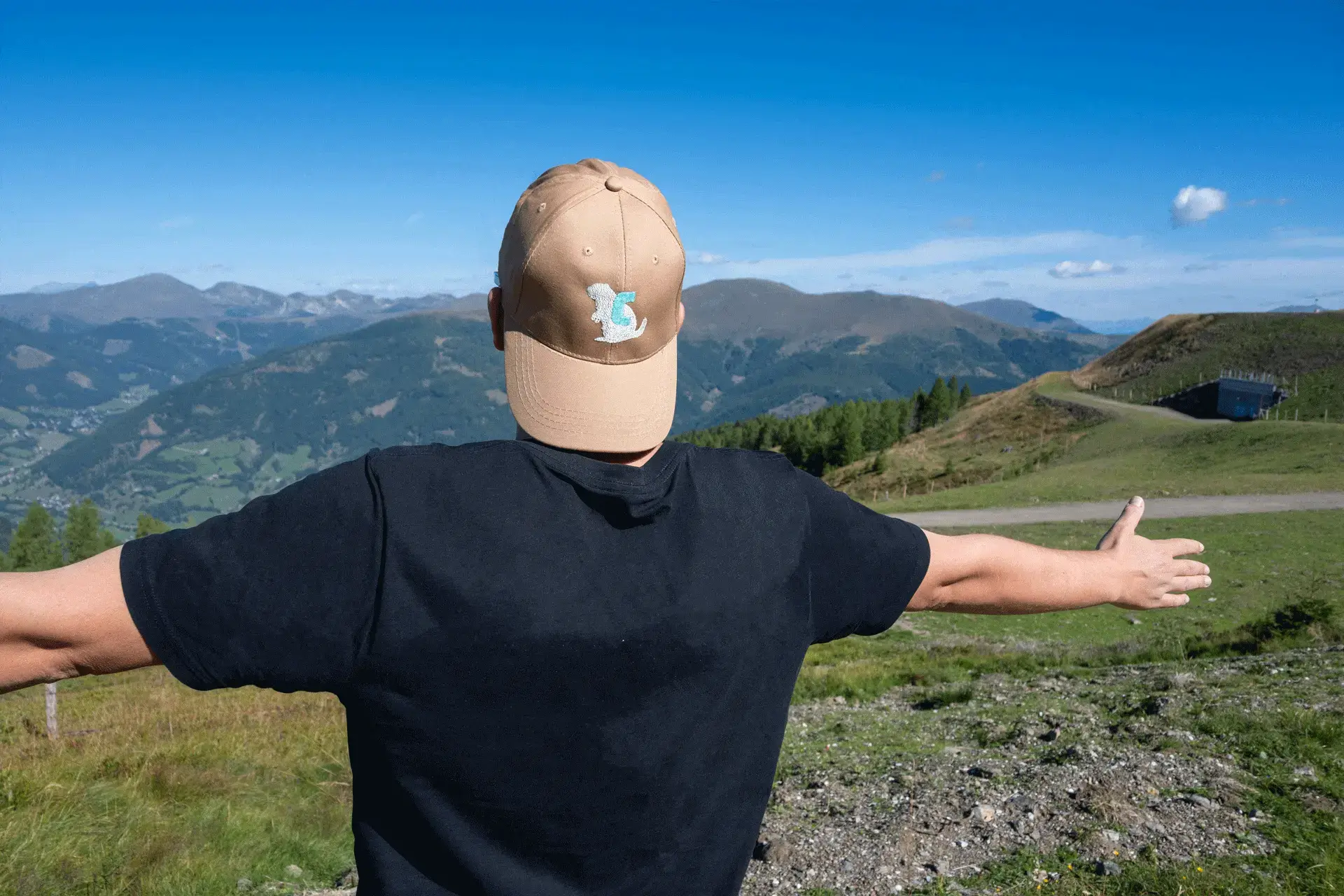 Person mit hellbrauner COOEE alpin Cap „Alinga“ breitet die Arme aus und genießt das Alpenpanorama – stylische Wanderkappe für Outdoor, Wandern und Urlaub in Österreich.
