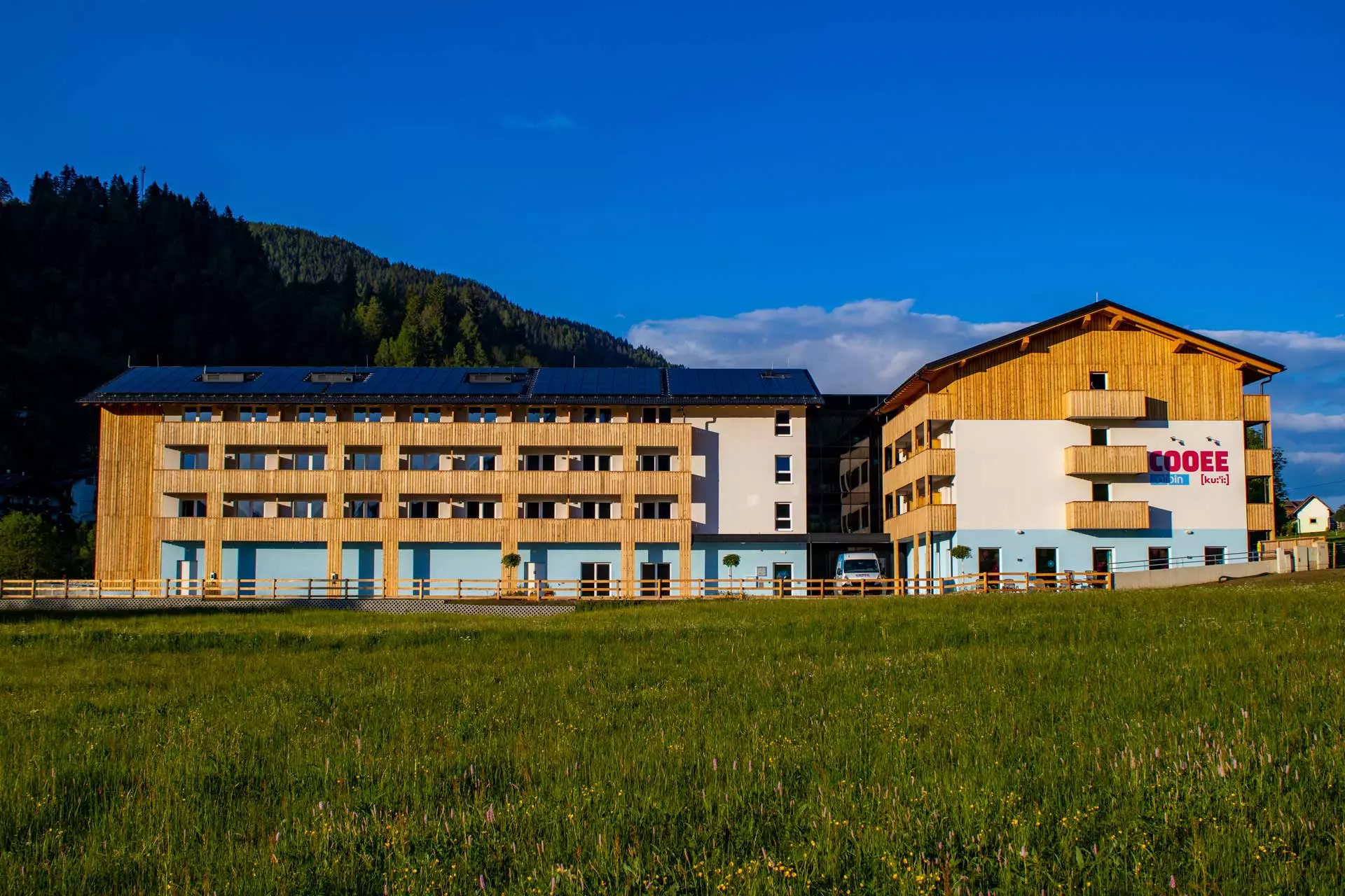 Das COOEE alpin Hotel Bad Kleinkirchheim von außen im Sommer. Im Vordergrund ist eine grüne Wiese und hinter dem Hotel sieht man auf einen mit Wald bedeckten Berg.