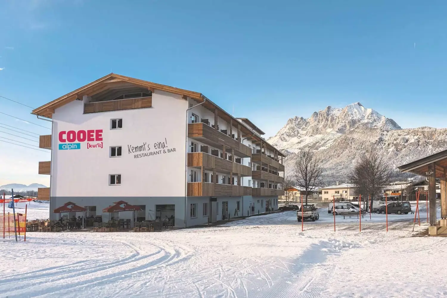 Außenansicht vom COOEE alpin Hotel Kitzbüheler Alpen im Winter. Vor der Sonnenterasse ist eine Spur durch den Schnee gezogen und im Hintergrund sieht man den verschneiten Wilden Kaiser.