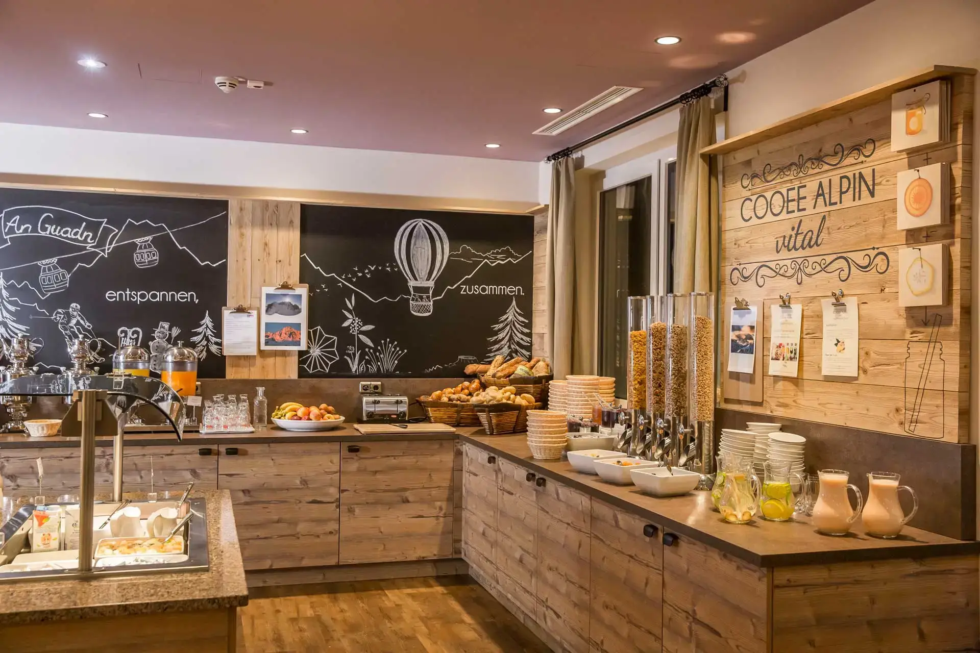 Un buffet dall'aspetto in legno su cui sono disposte brocche con acqua e limoni, muesli, pasticcini, cestini del pane, marmellata, frutta, un tostapane e un distributore di succo. A sinistra c'è un'isola buffet con yogurt e latte.