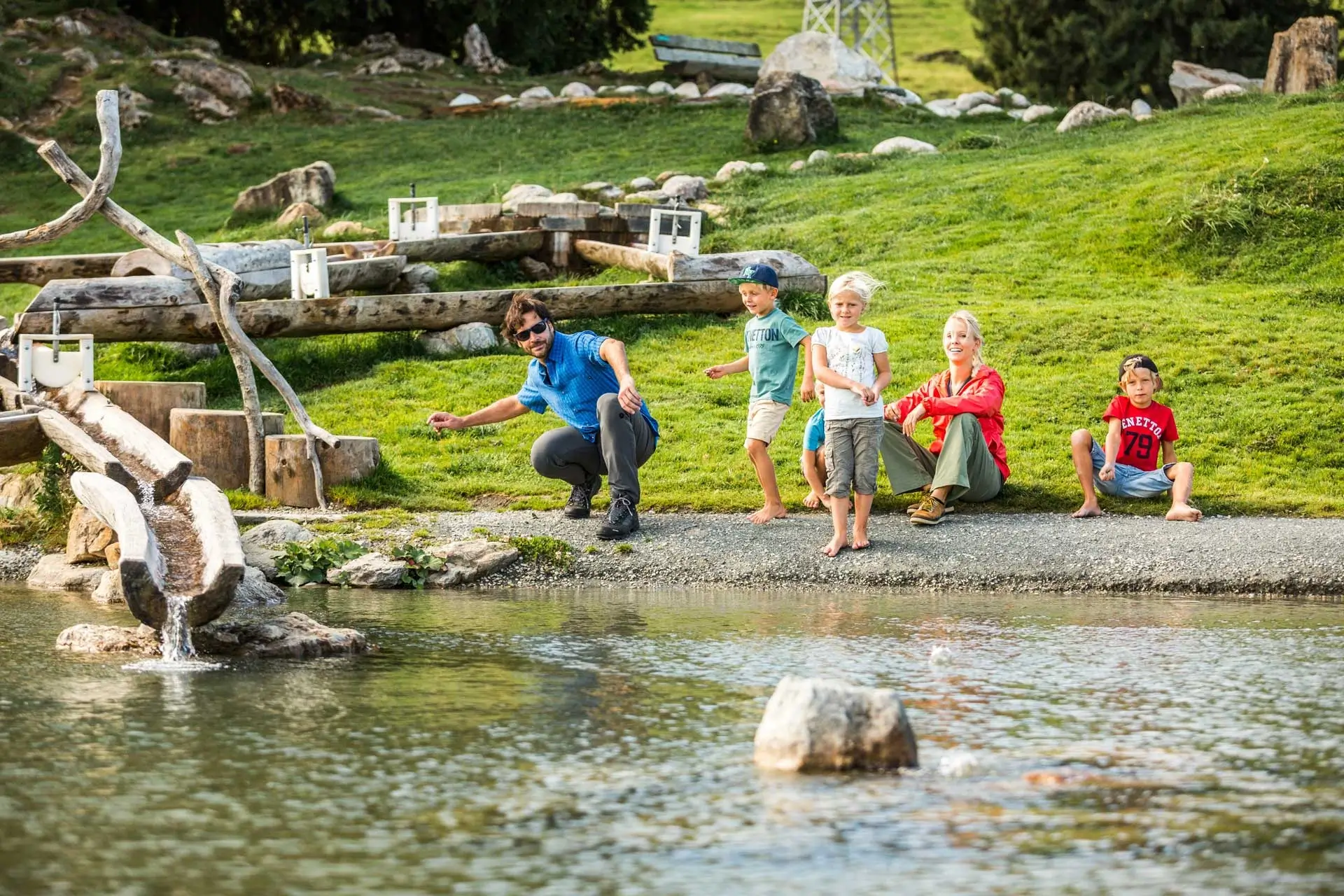Eine Familie sitzt am Wasser und genießt die Sonne. Der Vater wirft gerade einen Stein in den Teich.