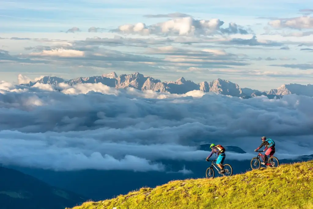 Zwei Personen mit Mountainbikes fahren einen Trail durch eine Bergwiese. Im Hintergrund ist ein beeindruckendes Bergpanorama und die Gipfel werden von der Sonne angestrahlt. Im Tal hängt eine Wolkendecke, aber am Berg scheint die Sonne.