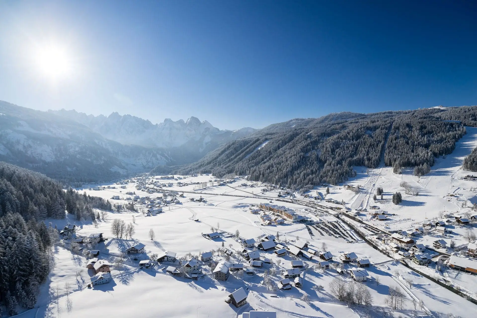 Eine Luftaufnahme von dem Gosautal im Winter. Man kann den Gosaukamm am Ende des Tals erkennen und links und rechts erstrecken sich Berge mit Wäldern und Skipisten. Im Tal ist der Ort Gosau mit seinen verschneiten Häusern.
