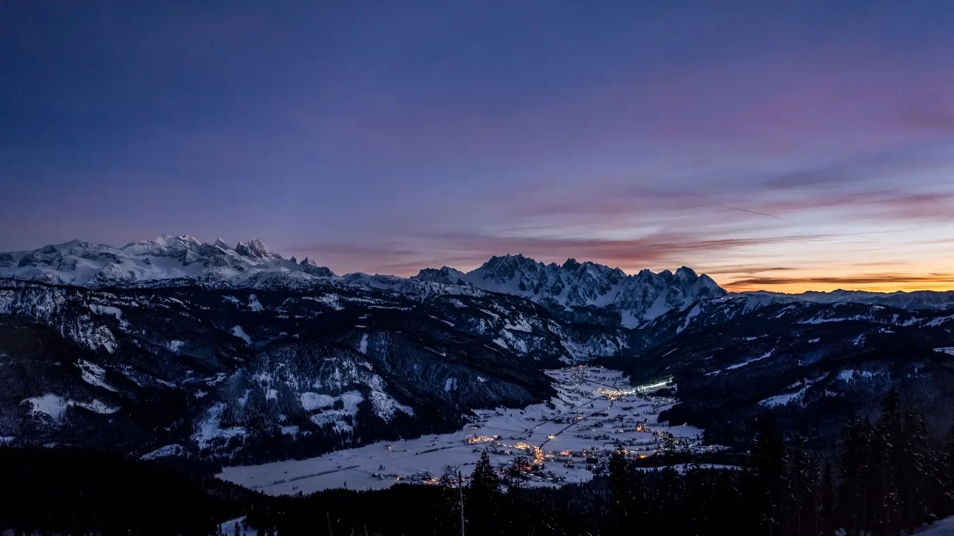 Panoramatický pohled na zasněžené údolí Gosau během modré hodiny s osvětlenými domy a výraznou horskou kulisou hřebene Gosaukamm.