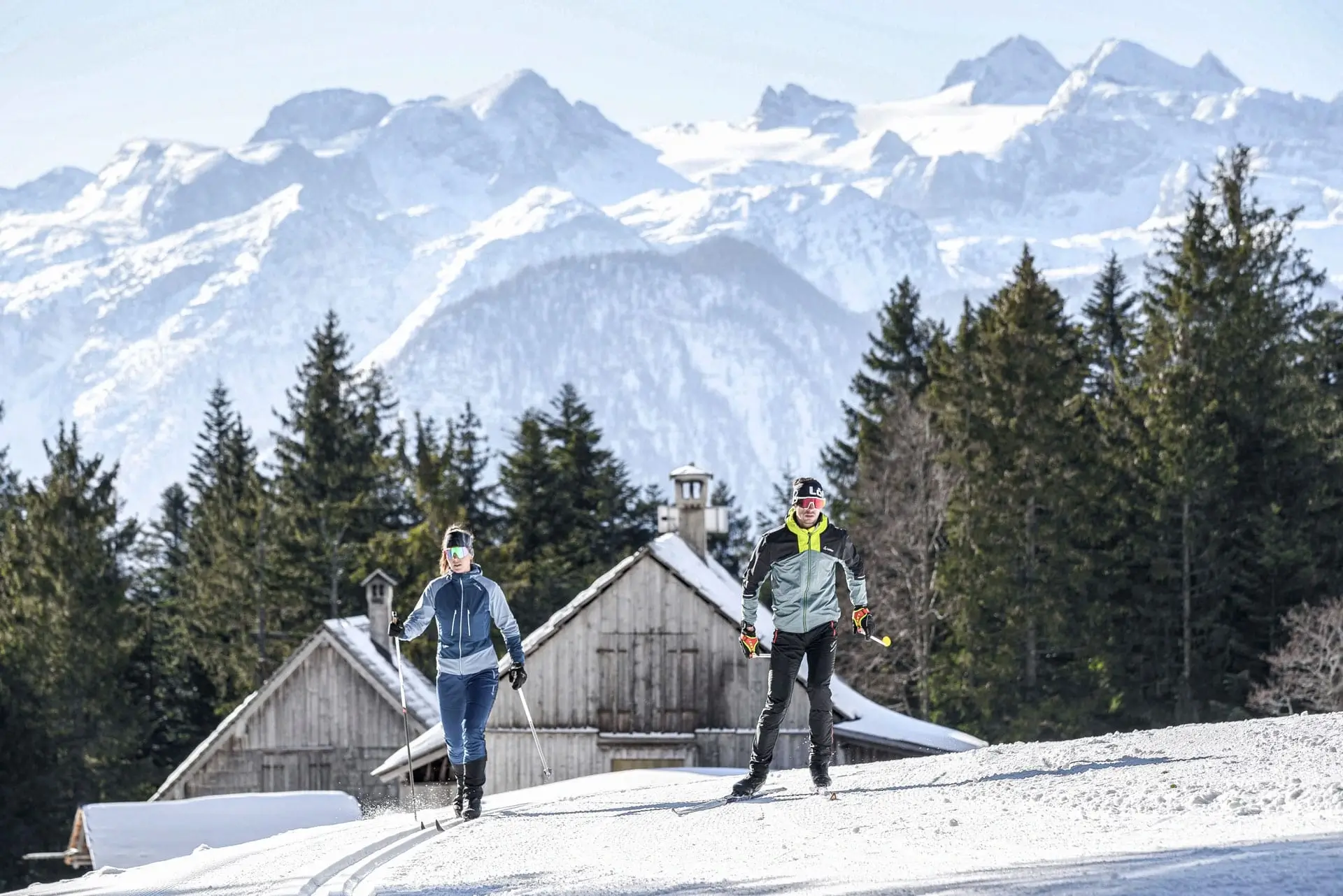 Due persone con gli sci di fondo: una segue la pista tracciata mentre l'altra pattina. Sullo sfondo si vedono una baita innevata, un bosco e un impressionante panorama montano.