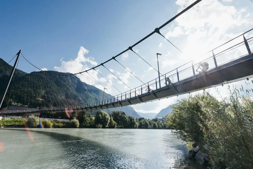 Drei Personen fahren mit Fahrrädern über eine lange Hängebrücke über die Drau. Das türkise Wasser ist bewegt und am Ufer wachsen große Bäume mit saftigen, grünen Blättern. Die Landschaft ist von Bergen umgeben und die Sonne scheint vom blauen Himmel herab.