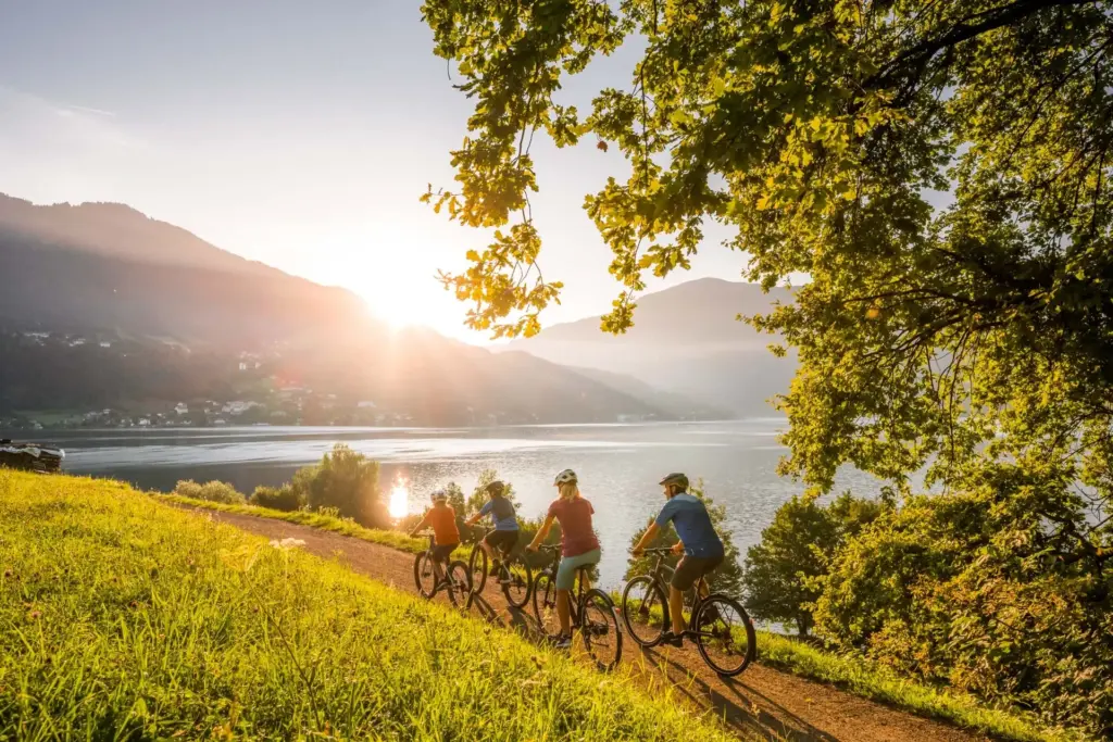 Vier Personen fahren mit Fahrrädern auf einem Radweg entlang des Ufers des Millstätter Sees. Im ruhigen Wasser des Sees spiegelt sich die Sonne die hinter dem dahinterliegenden Berg hervor scheint. Sie versetzt das Bild in eine warme Farbe und auf der gegenüberliegenden Uferseite ist ein kleines Bergdorf.