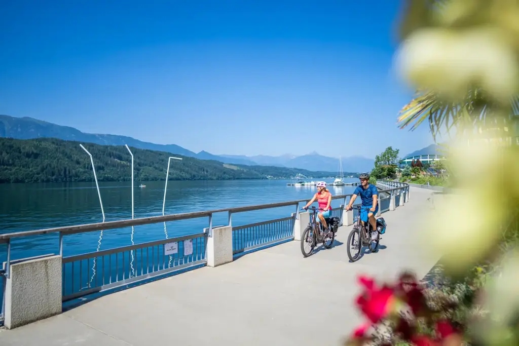 Zwei Personen fahren mit Fahrrädern einen asphaltierten Radweg am Millstätter See entlang. Das Ufer ist durch einen Zaun vom See abgetrennt und am Ufer wachsen bunte Blumen und grüne Bäume und Sträucher. Das Wasser des Millstätter Sees ist tief blau gefärbt und der Himmel ist wolkenlos an einem sonnigen Sommertag. Hinter den Radfahrern liegt ein kleiner Steg an dem drei weiße Boote anlegen. Auf der gegenüberliegenden Uferseite sind mit Wald bedeckte Berge zu erkennen die sich entlang des Sees in die Ferne erstrecken.