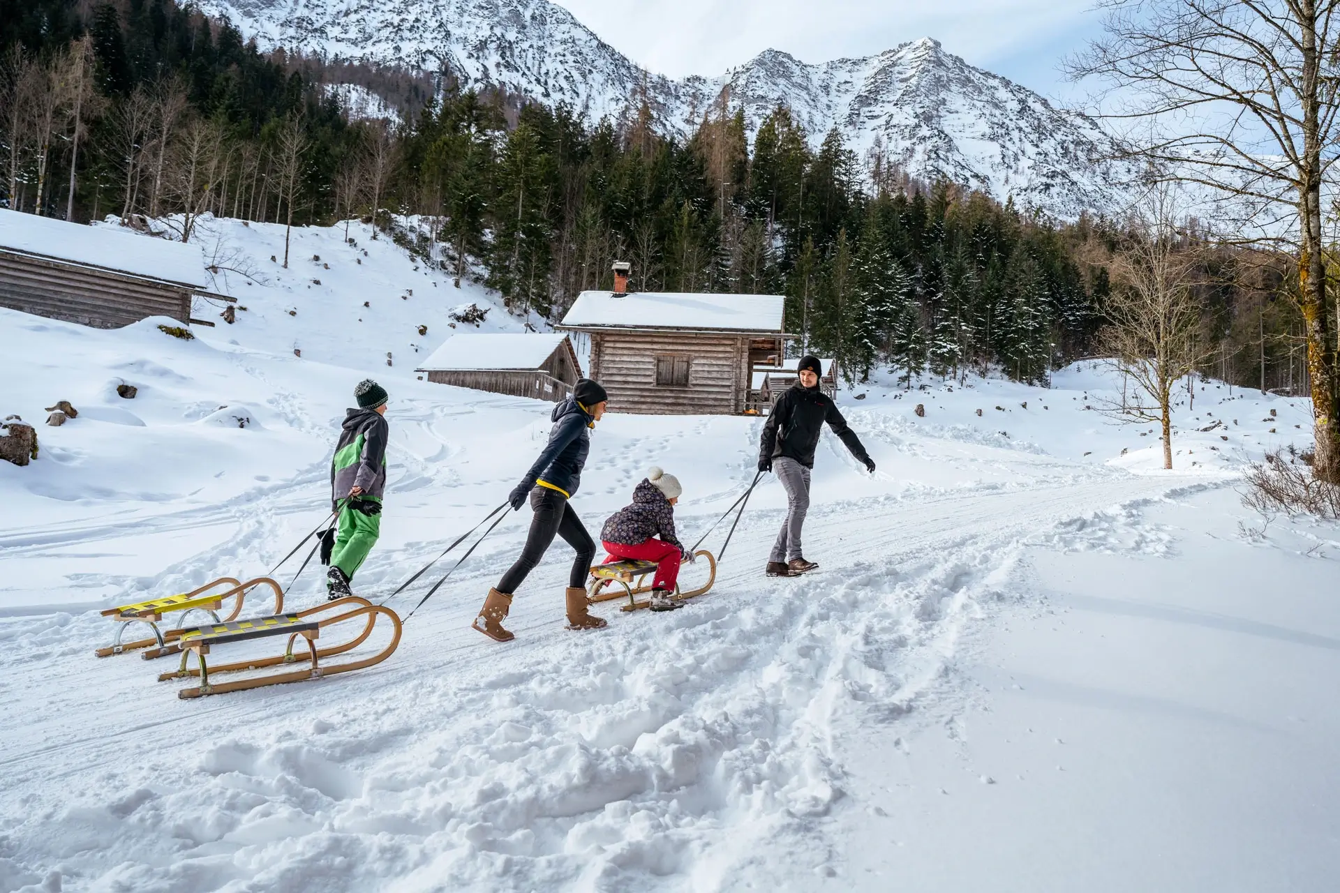 Tre persone tirano le slitte lungo un sentiero innevato. Un bambino siede su quella anteriore e viene trascinato. Il sentiero passa accanto a una capanna e si inoltra in una foresta. Sullo sfondo si vedono montagne innevate.