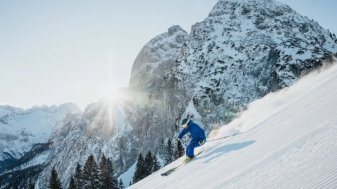 一位身着蓝色滑雪服的滑雪者正在滑雪道上转弯。阳光从白雪覆盖的群山后面照射出来。