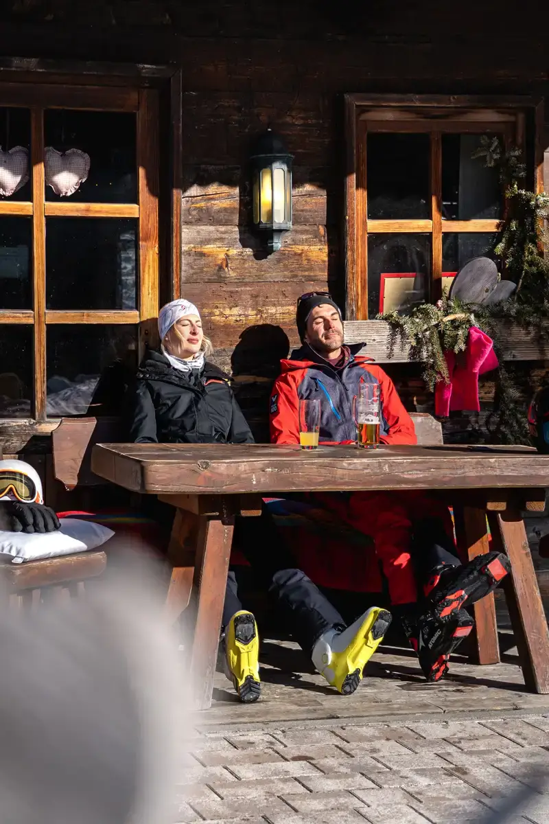 Zwei Personen in Skikleidung entspannen an einem Holztisch auf der Sonnenterrasse einer Hütte mit zwei Getränken und genießen mit geschlossenen Augen die Sonne in ihren Gesichtern.