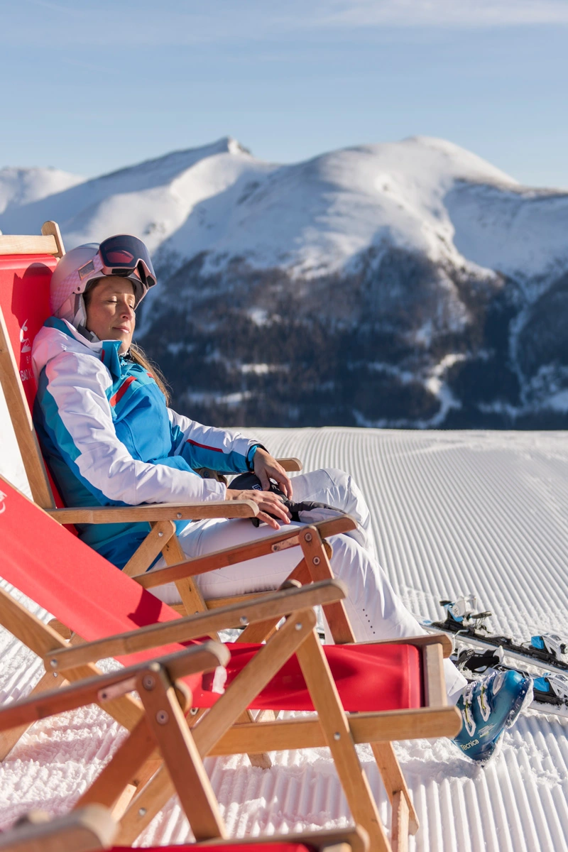 Eine Person in Skikleidung sitzt in einem roten Liegestuhl auf einem Berg und genießt mit geschlossenen Augen die Sonne in ihrem Gesicht. Vor ihr liegen Ski und im Hintergrund blickt man auf ein verschneites Bergpanorama.