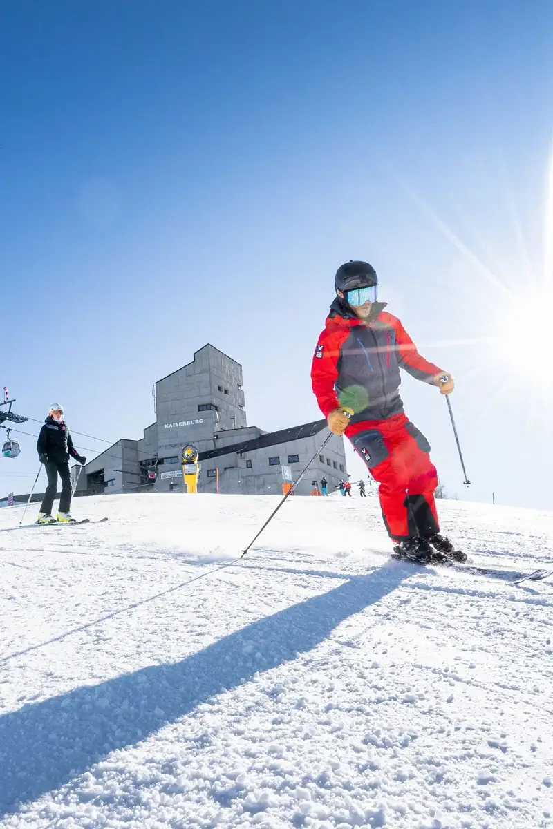 Two people are skiing one behind the other on a slope. Behind them, the sun shines down from the blue sky and the mountain station of a cable car can be seen.