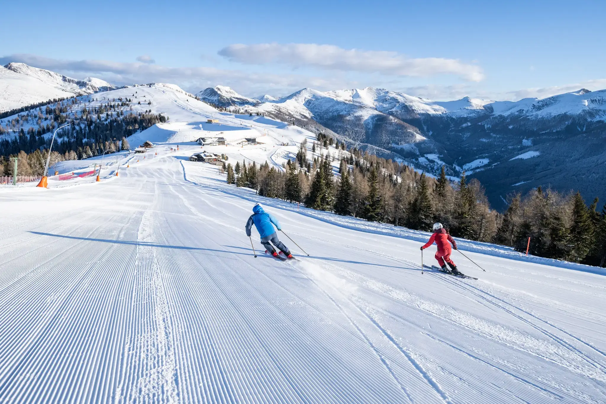 Zwei Skifahrer:innen fahren bei Sonnenschein auf einer frisch präparierten Piste in den österreichischen Alpen, umgeben von verschneiten Bergen und Wäldern – Skiurlaub mit Panoramablick in Bad Kleinkirchheim.