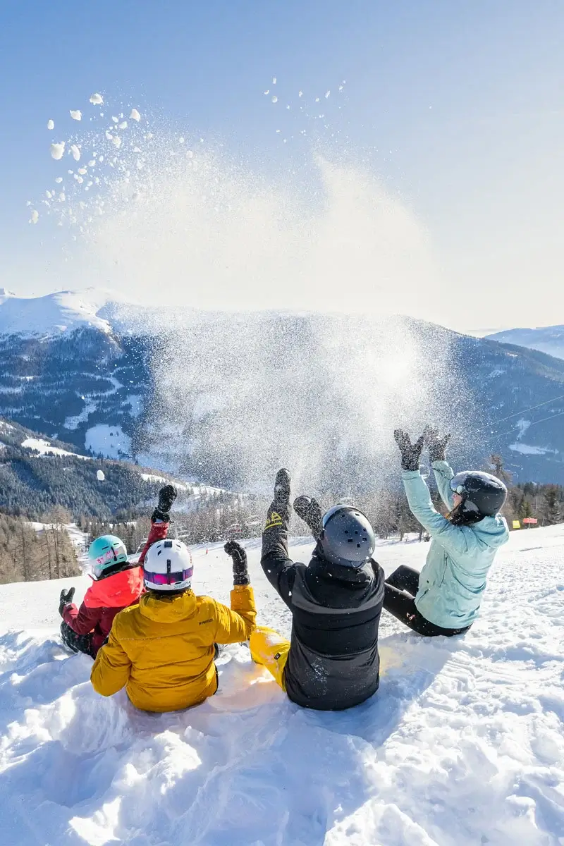 Four people in ski clothing sit in the snow on a mountain and throw snow into the air. You can see from the snow-covered mountain peaks down into the valley.