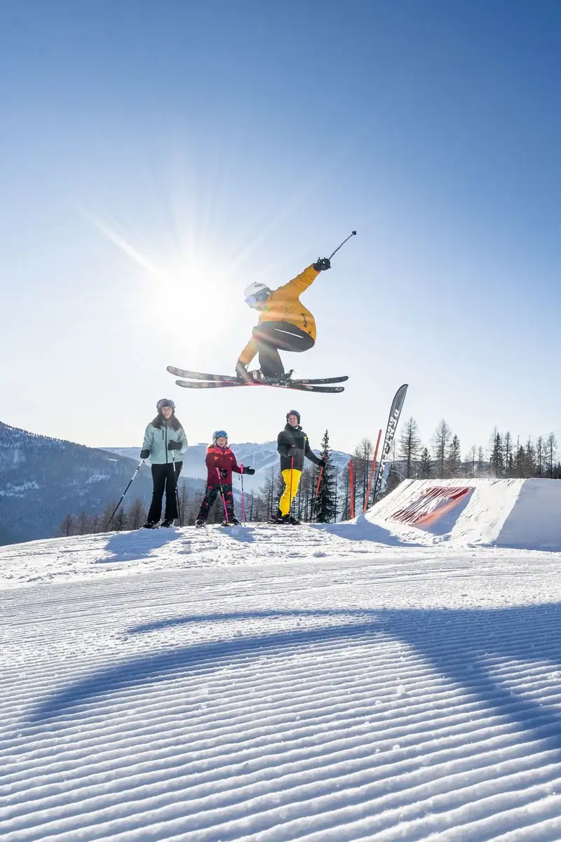 Three people in ski clothing watch a person jumping over a ski jump on skis. The sun is shining down from the blue sky and the slope is perfectly groomed.