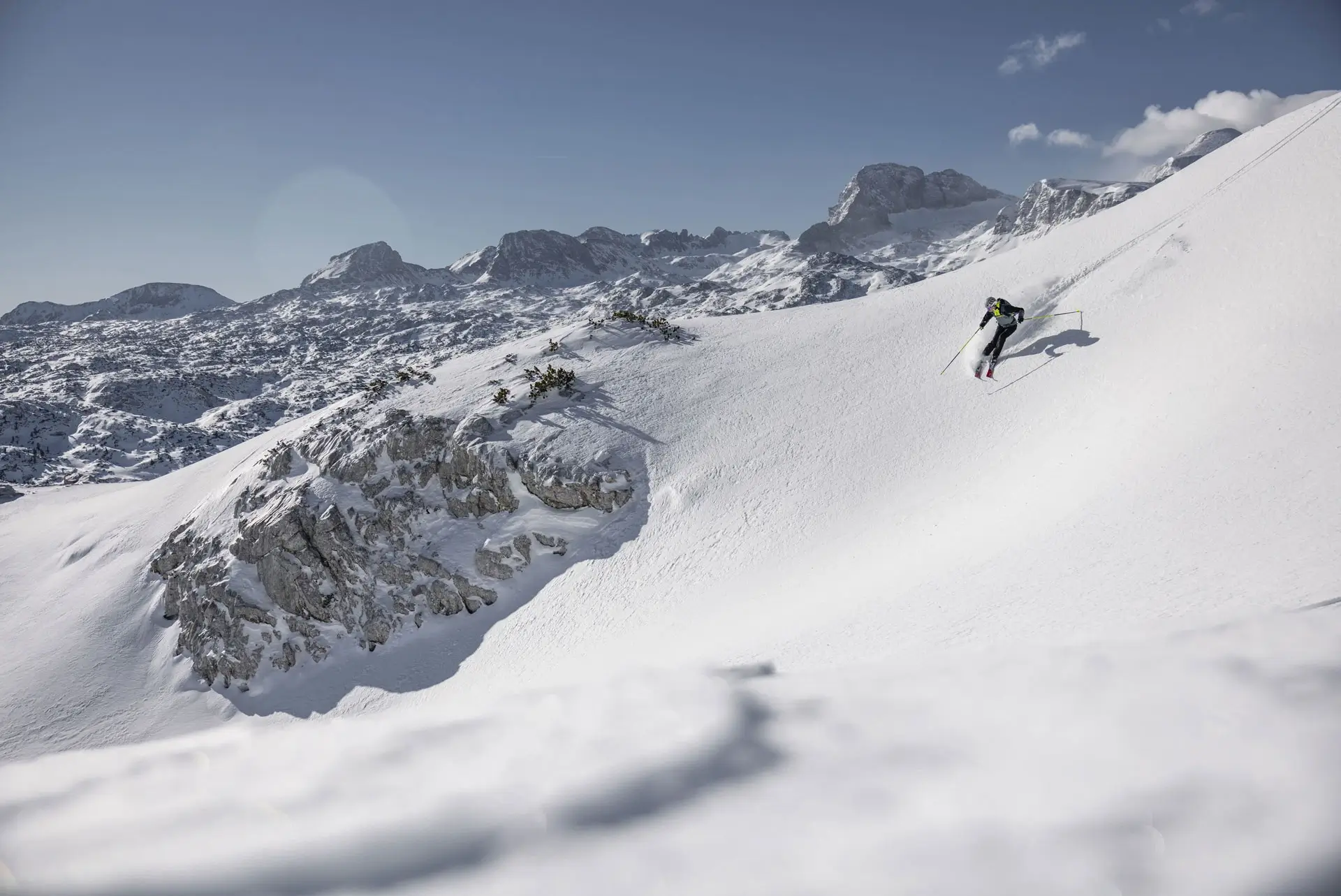 Uno sciatore sta sciando su una pista intatta con neve alta. Sullo sfondo si vede un panorama montano con rocce e piccoli alberi.