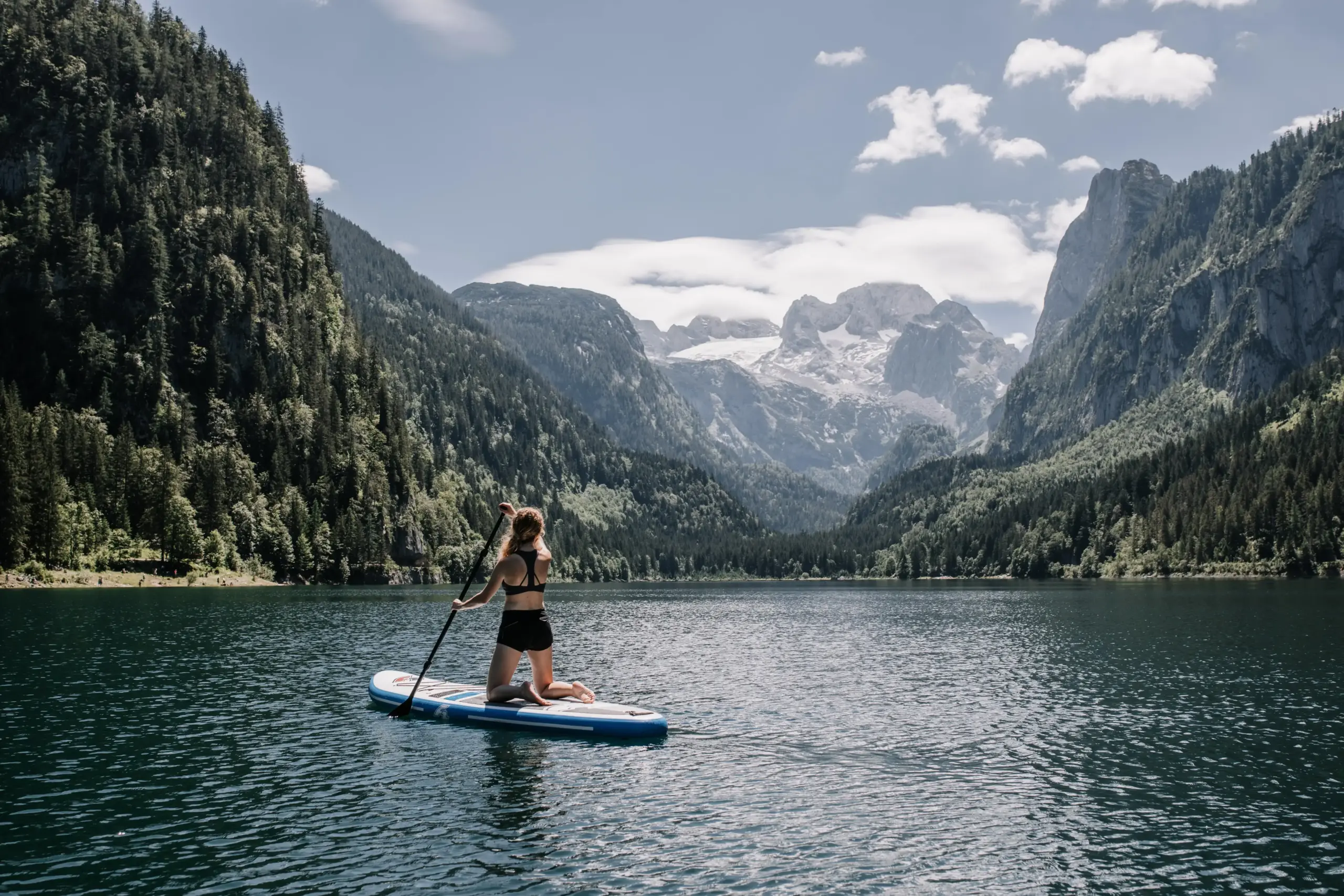 Eine Person mit schwarzer Badekleidung und lagen Haaren kniet auf einem Stand-Up-Board auf einem Bergsee und taucht mit einem Paddel an. Das Wetter ist sonnig und sie blickt auf ein beeindruckendes Bergpanorama. Die Berge sind mit grünen Wäldern bedeckt und auf den Gipfeln liegt weißer Schnee.