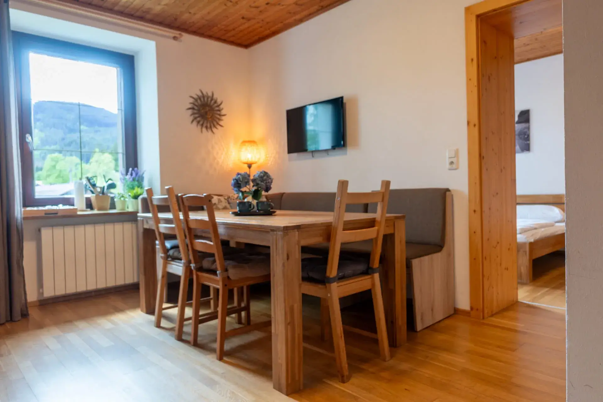Útulný obývací a jídelní kout v apartmánu u jezera Traunsee v komplexu Ulmenhof v Gosau s velkým jídelním stolem, lavicí, televizí a výhledem do zeleně.