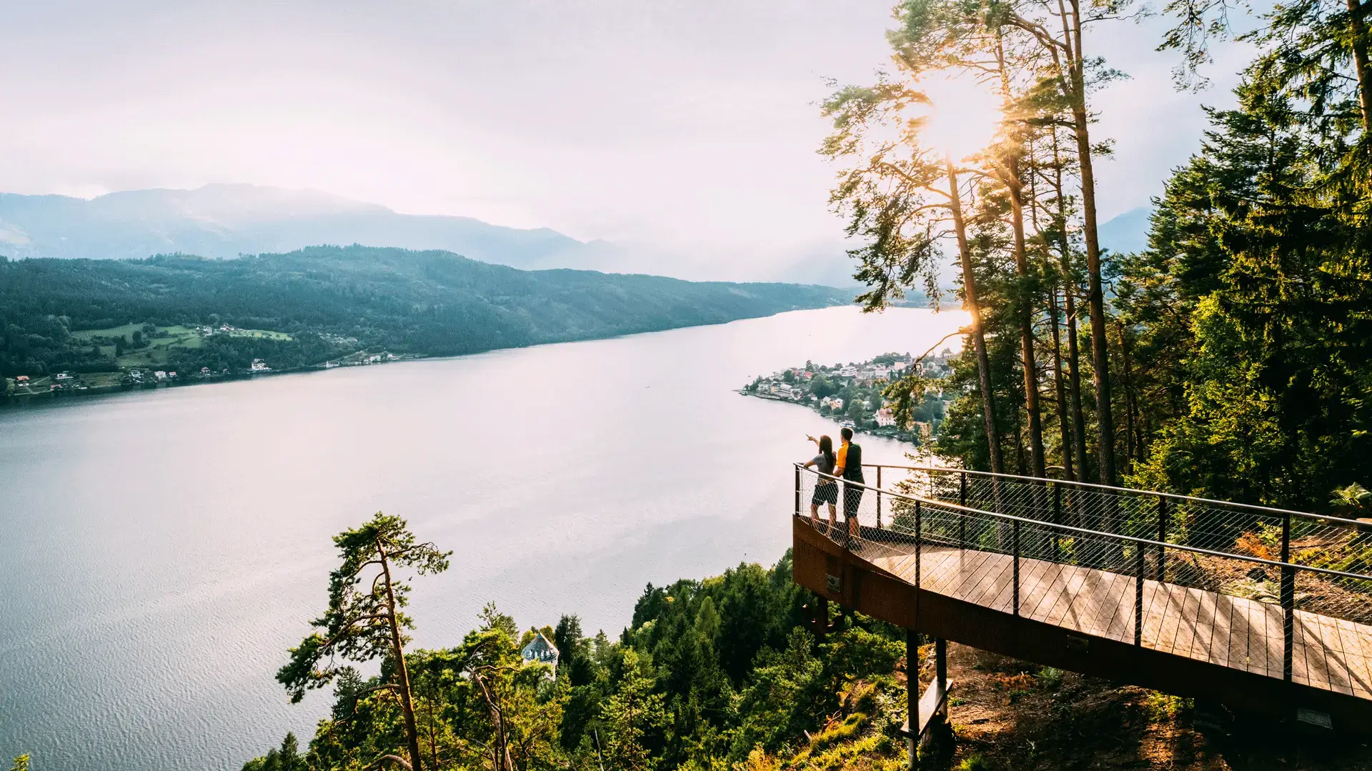 Aussichtspunkt mit Holzplattform über dem Millstätter See, umgeben von Wald und Bergen bei Sonnenlicht.