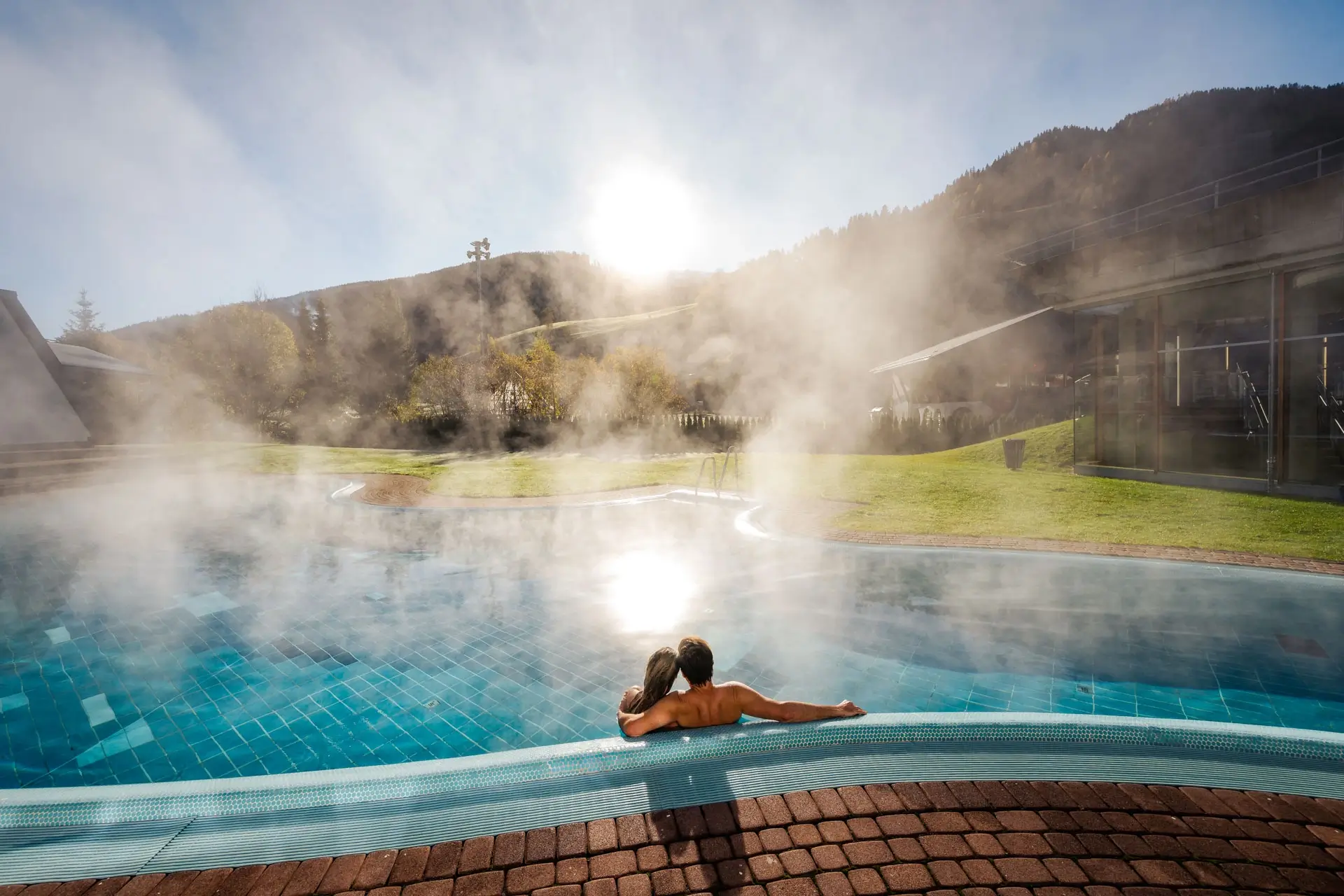 Zwei Personen liegen sich in einem Außenbecken einer Therme in den Armen. Das Wasser dampft und Gelb verfärbte Bäume bedecken die umliegenden Berge.