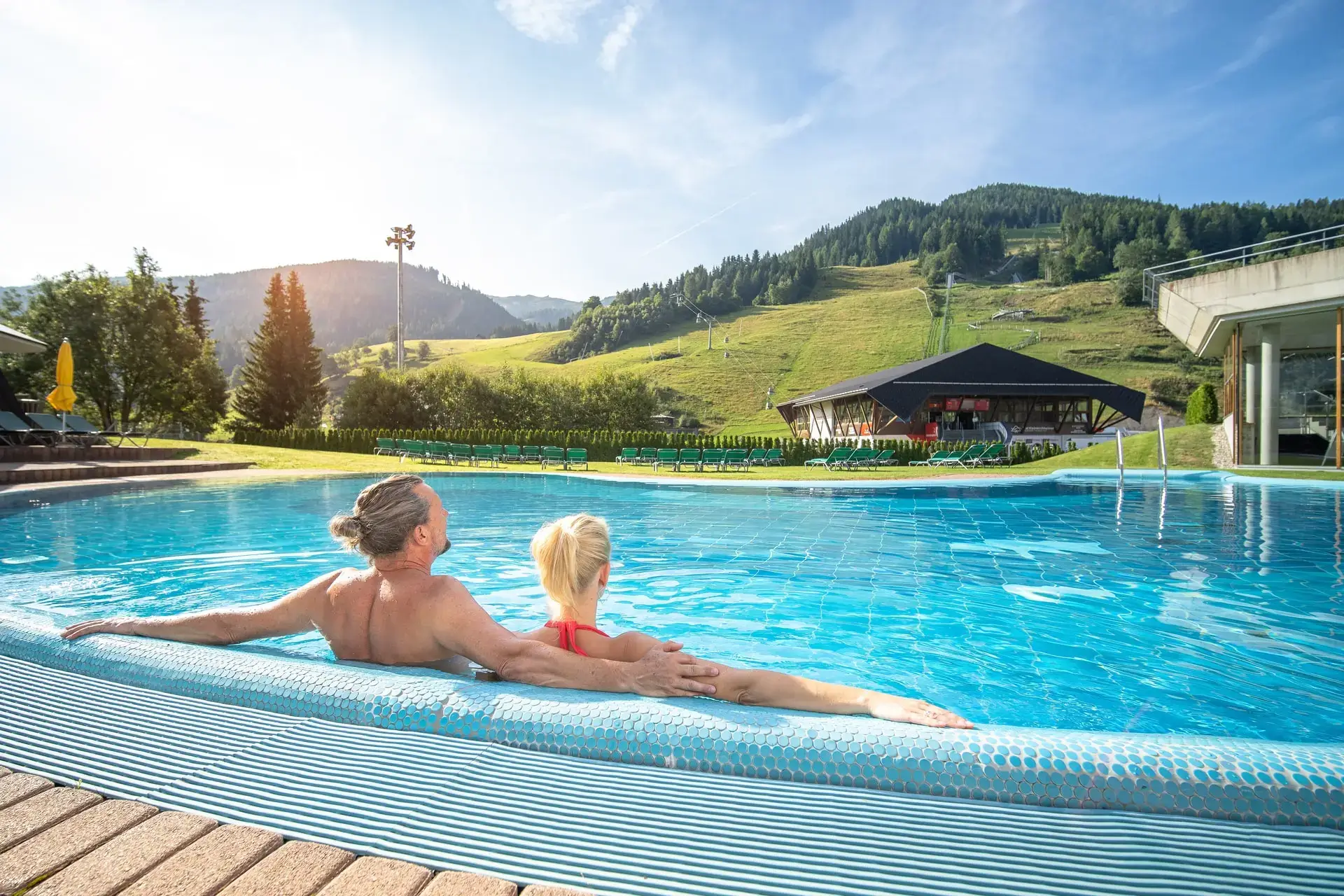 Zwei Personen sind nebeneinander in einem Außenbecken einer Therme. Der Ausblick ist auf grüne Wiesen, Wälder, Berge und eine Bergbahn. Neben dem Becken stehen grüne Liegestühle auf der Wiese.