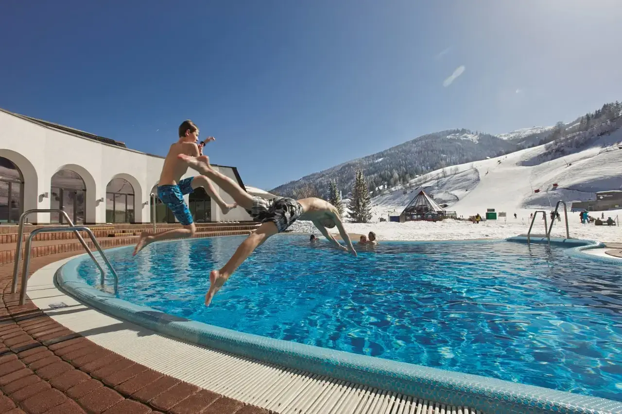Zwei Personen springen in Badekleidung in das Außenbecken einer Therme. Hinter dem Becken sind schneebedeckte Berge und eine Skipiste.