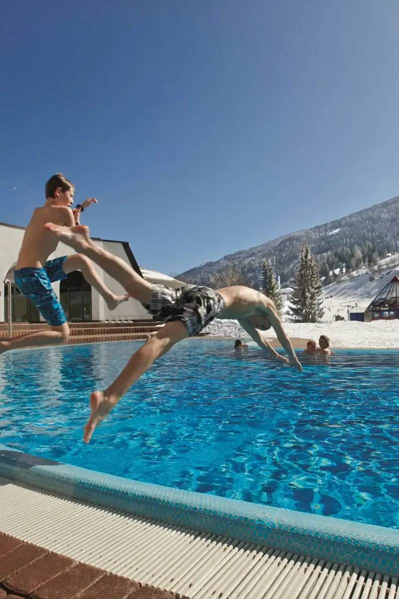 Two people in bathing suits jump into the outdoor pool of a thermal spa. Behind the pool are snow-covered mountains and a ski slope.